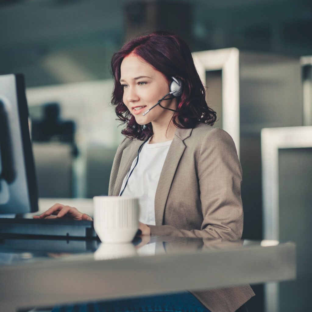 Beautiful young woman telephone operator with headset working on desktop computer in customer service call support helpline business center with teamworker in background
